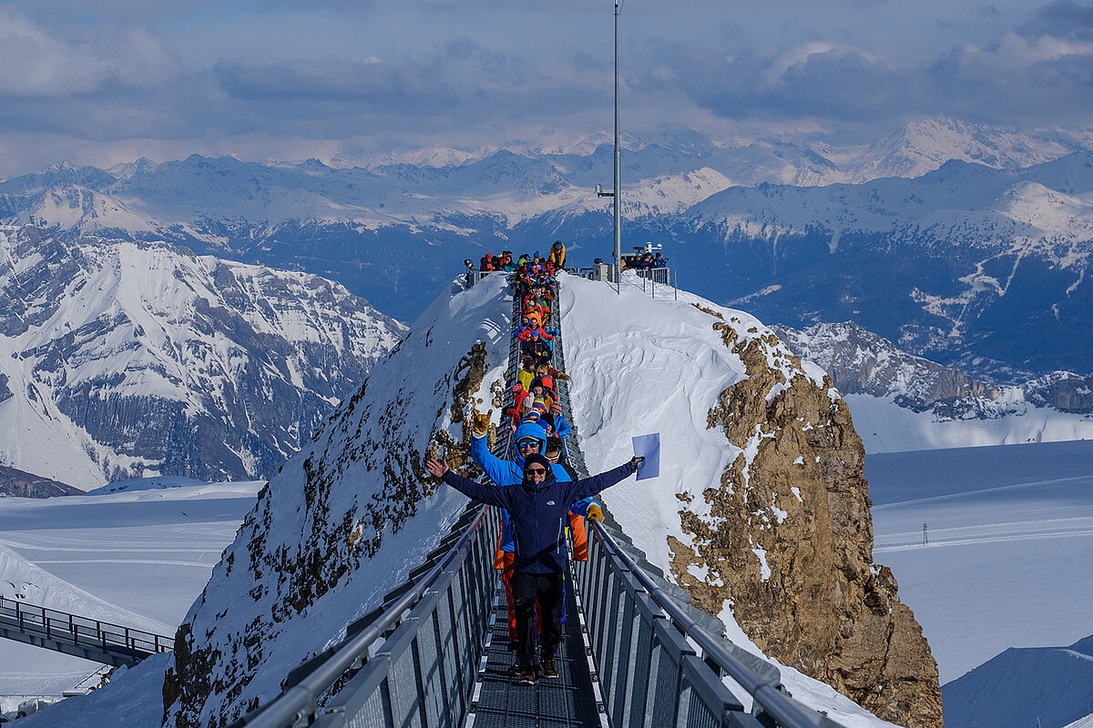 Winter hiking is very popular in Glacier3000. People in winter clothes walk across a snowy bridge.
