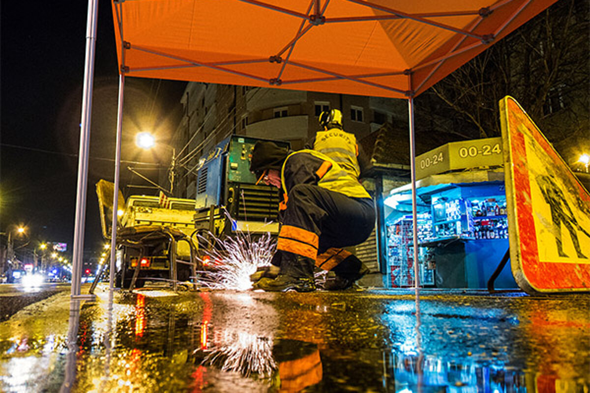Pro-Tent Bauzelte halten allen extremen Bedingungen auf Baustellen stand. Ein Bauarbeiter kniet auf einer Strasse unter einem Montagezelt und arbeitet mit einer Flex.