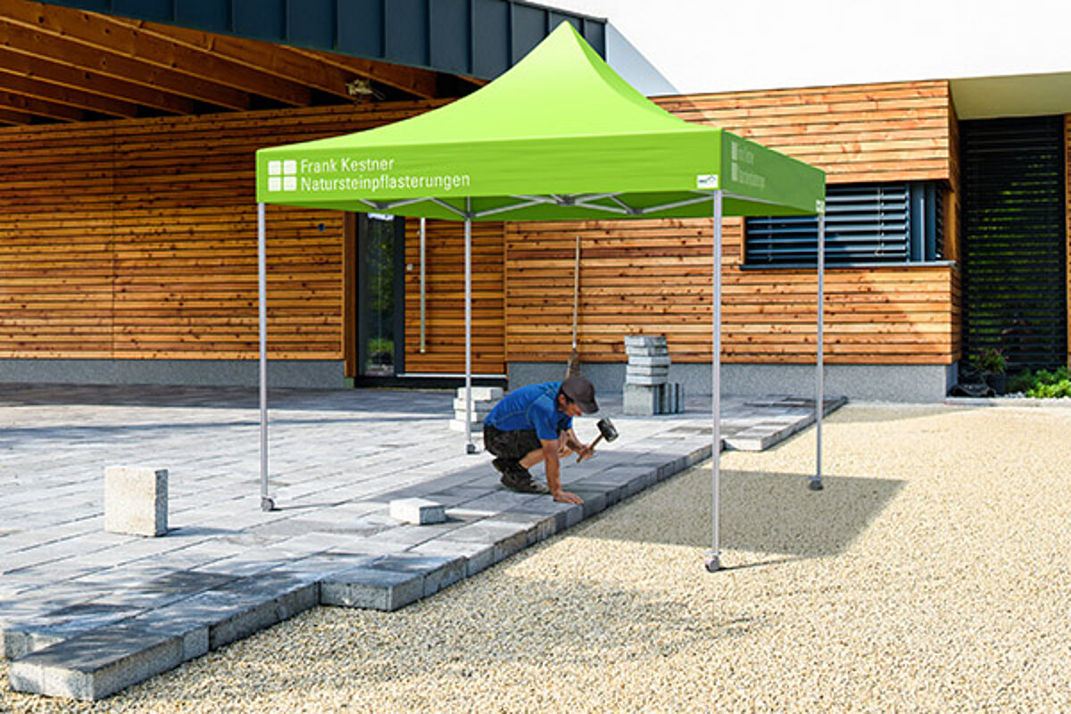 Unsere Arbeitszelte sind ideal für Handwerker, die draussen arbeiten! Ein Bauarbeiter verlegt eine Terrasse im Schatten, den ihm ein Pro-Tent Bauzelt spendet.