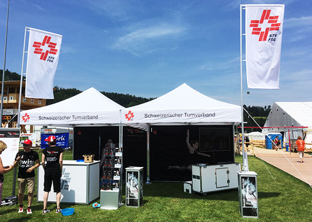 We can offer you accessories such as flags and counters for your club tent. A club tent hosted by the Swiss Gymnastics Federation stands in a meadow.
