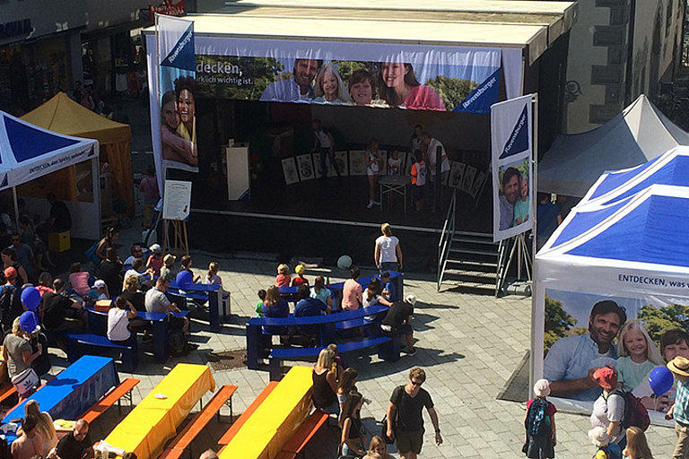 A Ravensburger event with a stage, folding tents and beer benches taking place on a public square.  